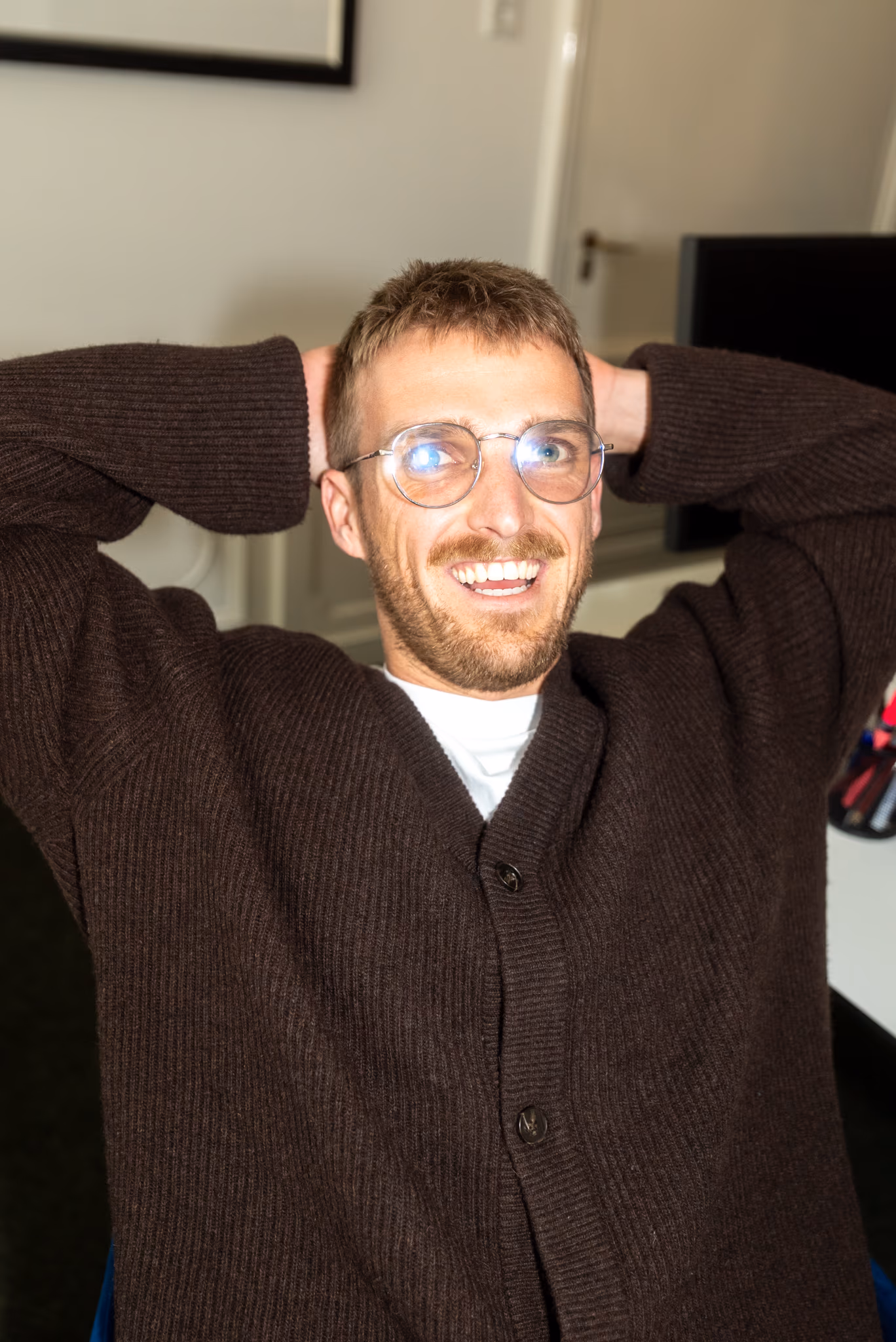 Homme souriant à lunettes en cardigan brun, mains derrière la tête