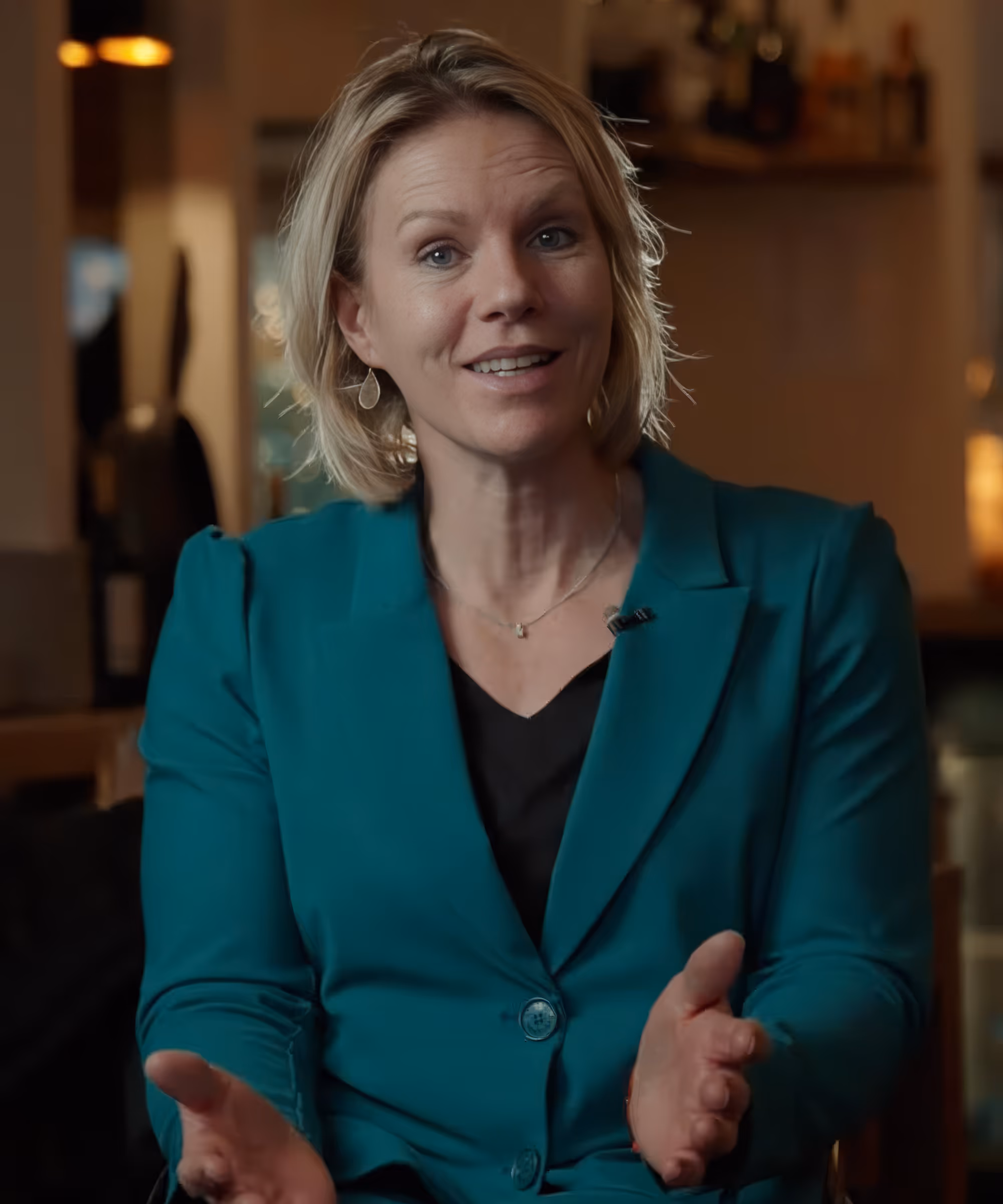 A smiling woman in a teal blazer speaks and gestures, wearing a lapel microphone
