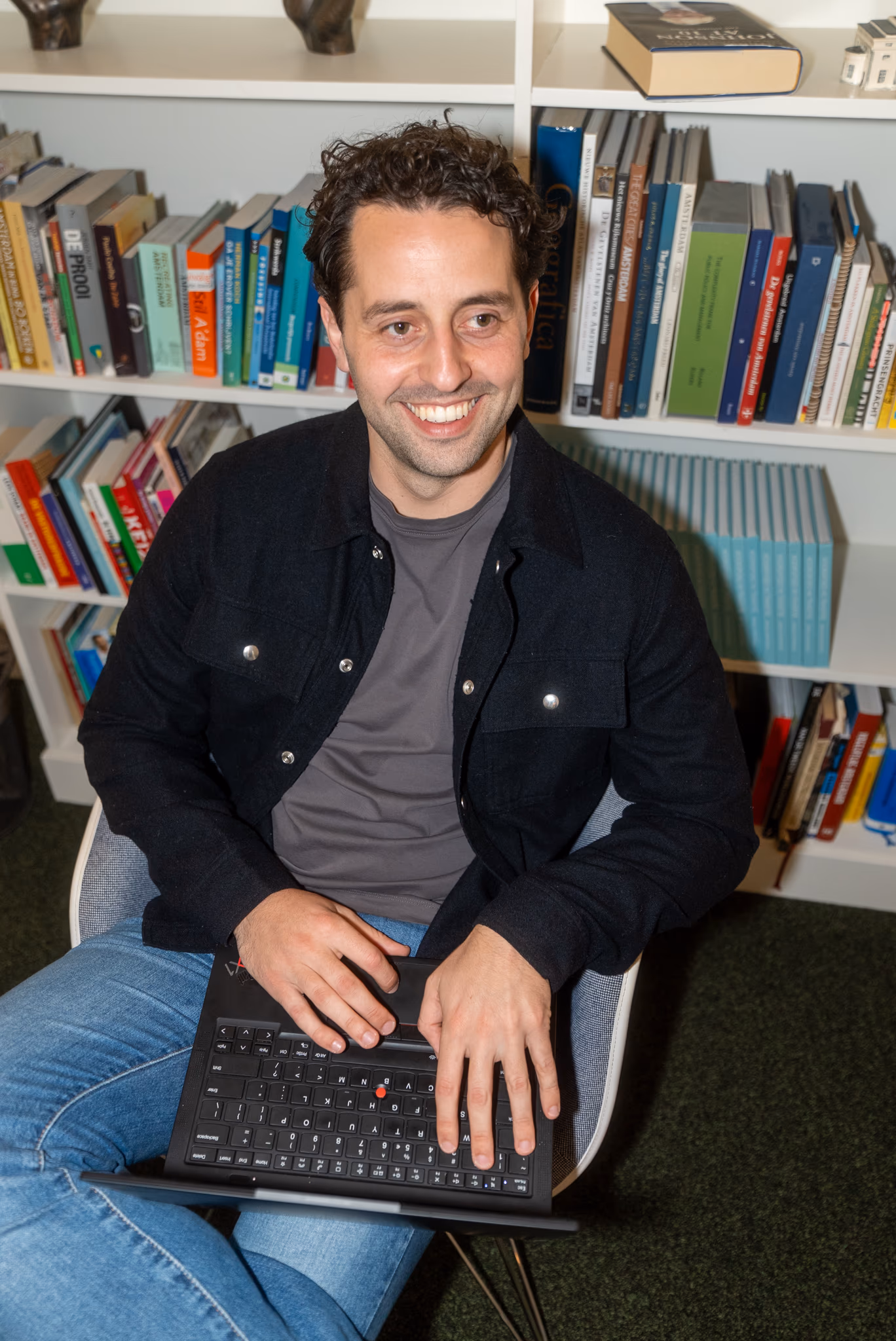 Lächelnder Mann mit lockigen Haaren sitzt mit einem Laptop im Sessel, vor einem vollen Bücherregal