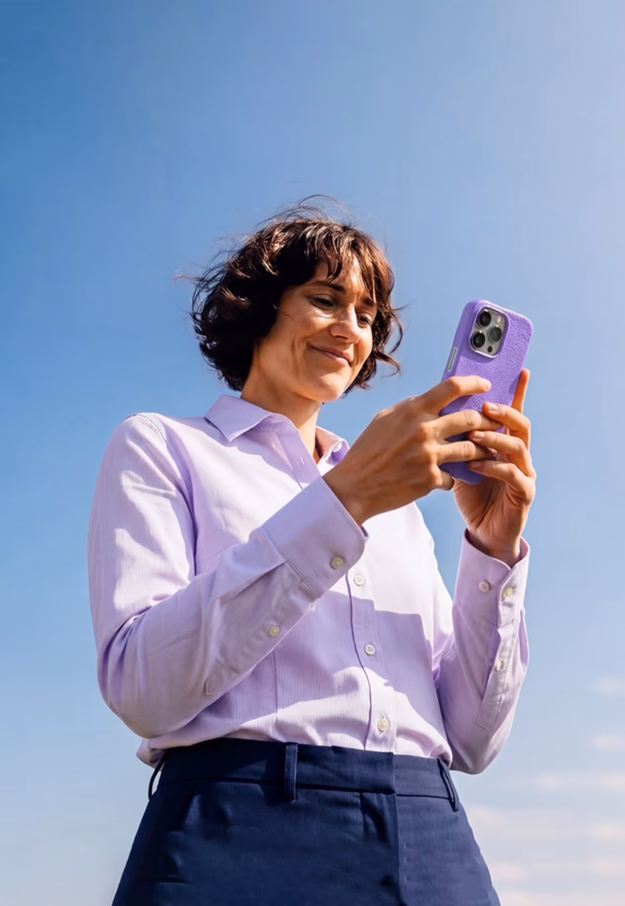 Woman in a lilac shirt holding a purple-cased phone against a blue sky